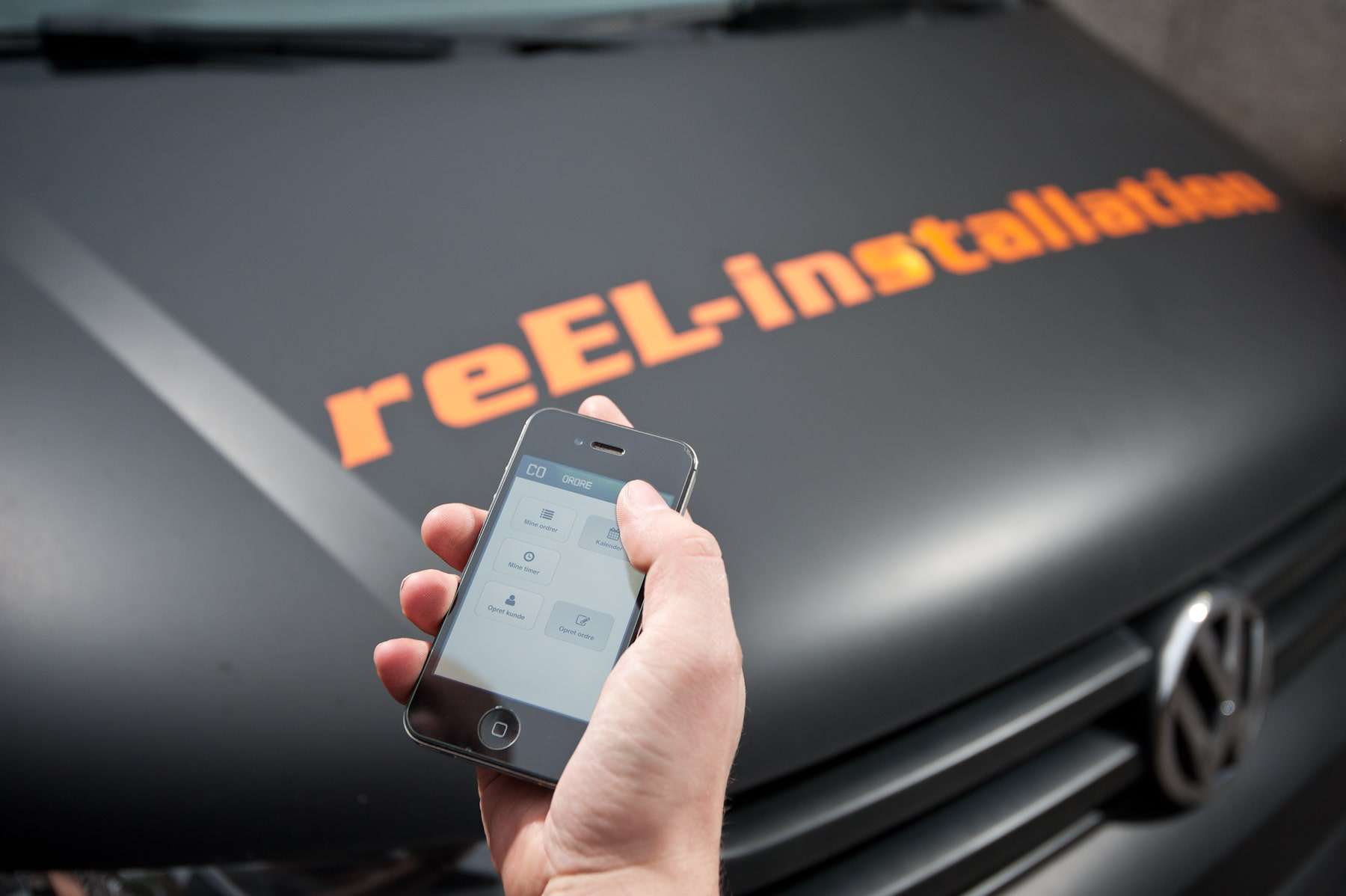 Mobiltelefon i hånden på en person og reEl-installation logo i baggrunden på en bil